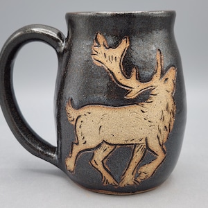 May include: A dark gray ceramic mug with a large handle. The mug features a tan-colored relief carving of a running elk. The elk is centered on the mug's body, with its antlers and body details clearly visible. The mug has a glossy finish.