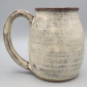 Może przedstawiać: Ceramiczny kubek o zaokrąglonym korpusie i dużym uchwycie. Kubek posiada nakrapiane, kremowobiałe szkliwo z ciemnobrązowymi plamkami. Krawędź oraz uchwyt są podkreślone ciemniejszym odcieniem brązu, co tworzy kontrast z jasnym korpusem naczynia.