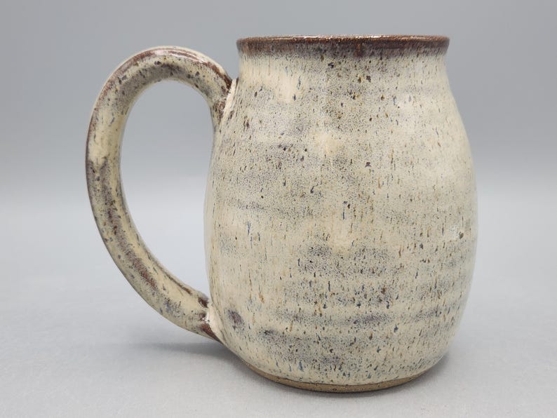 Może przedstawiać: Ceramiczny kubek o zaokrąglonym korpusie i dużym uchwycie. Kubek posiada nakrapiane, kremowobiałe szkliwo z ciemnobrązowymi plamkami. Krawędź oraz uchwyt są podkreślone ciemniejszym odcieniem brązu, co tworzy kontrast z jasnym korpusem naczynia.