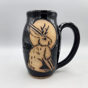 Może przedstawiać: Czarny ceramiczny kubek z beżowym reliefem przedstawiającym jackalope z długimi uszami i rogami. Stworzenie jest wyśrodkowane na okrągłym beżowym tle, a kubek posiada zakrzywione ucho po prawej stronie.