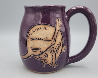 May include: A purple ceramic mug with a speckled finish and a large handle. The front features a carved, light tan illustration of a cat in a curled position with one leg extended upward, showcasing detailed line work and a smooth glaze.