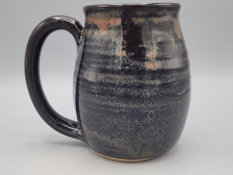 Może przedstawiać: Ceramiczny kubek o zaokrąglonym korpusie i dużym uchwycie. Powierzchnia pokryta jest ciemnym, błyszczącym szkliwem w odcieniach czerni i głębokiego błękitu, podkreślonym subtelnymi poziomymi żłobieniami oraz jasnobrązowym rantem.