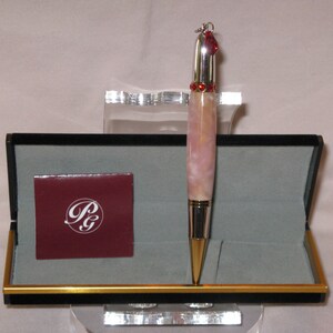 May include: A pink and silver pen with a red heart charm and rhinestones. The pen is in a black box with a gold trim and a red label with the letters "P&G".