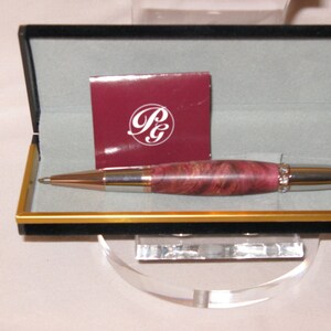 May include: A wooden pen with a pink and brown swirl pattern, silver accents, and a crystal charm. The pen is in a black velvet box with a gold trim. The box also contains a small red card with the letters "P&G" in a circle.