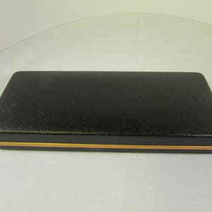 Puede incluir: Una caja rectangular negra con un borde dorado. La caja está hecha de un material texturizado y tiene una parte superior lisa y plana.