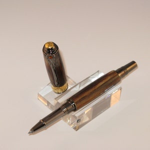 Könnte beinhalten: Ein brauner und silberner Stift mit goldener Verzierung und einem transparenten Acrylständer. Der Stift hat eine Holzmaserung und ein kleines orangefarbenes Juwel oben.