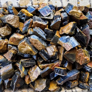 Puede incluir: Primer plano de un montón de piedras de ojo de tigre en bruto y talladas toscamente. Las piedras son una mezcla de colores marrón, dorado y azul.