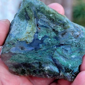 Puede incluir: Una pieza grande de ágata verde musgo de forma irregular con un centro de ágata azul y blanco. El ágata tiene una variedad de texturas y patrones, incluyendo remolinos, bandas y manchas.