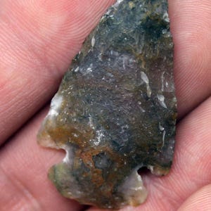 May include: A hand-held, ancient arrowhead. The arrowhead is made of stone with a mottled pattern of green, brown, and white. The arrowhead has a notched base and a sharp point.