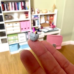 May include: A small grey cat figurine with pink ears and a pink tail. The cat is holding a pencil and a pink rubber in its paws. The figurine is sitting on a finger.