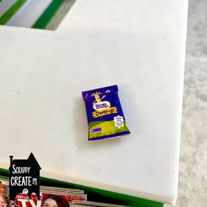 May include: A miniature packet of "String & Things Cheesestrings" cheese snacks. The bag is dark blue with yellow and green details. The product name and a giraffe cartoon are visible on the front.