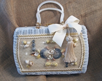 Könnte beinhalten: Hellblaue Korbtasche mit transparenten Kunststoffgriffen, verziert mit Muscheln, einer cremefarbenen Schleife und einer Quaste. Die Tasche hat einen goldenen Besatz und eine Spitzentafel.