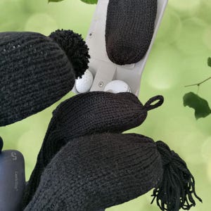 May include: Four black knitted golf club head covers with pom poms. Two covers are in a golf club holder with two golf balls. The holder has the text "Callaway" on it.