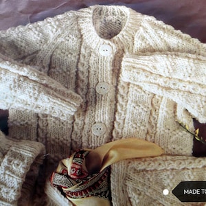 May include: A white knitted cardigan with a cable knit pattern and button closure. The cardigan is made to order.