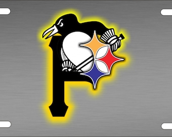 May include: A black and white Pittsburgh Penguins logo with a yellow outline on a silver metal license plate. The logo features a penguin with a hockey stick and a star with the colors of the Pittsburgh Penguins.