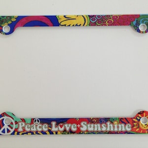 May include: A colorful license plate frame with a psychedelic design. The frame features a peace sign, a heart, and the text "Peace Love Sunshine".