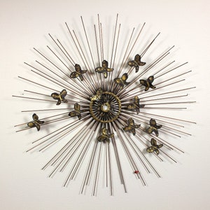 May include: A gold metal wall sculpture with a sunburst design and butterfly accents. The sculpture has a circular center with radiating spokes and multiple butterfly shapes.