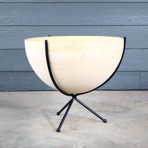 May include: A white ceramic planter with a rounded shape sits on a black metal stand with three legs. The stand is made of thin metal and has a simple design.