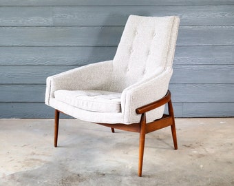 May include: A white upholstered armchair with a tufted back and wooden legs. The chair is sitting on a concrete floor in front of a gray wood wall.