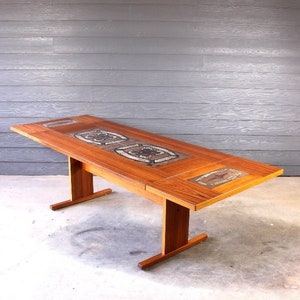 May include: A mid-century modern teak dining table with a tiled top featuring a geometric pattern. The table has a single pedestal base with a rectangular top.