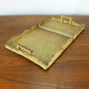May include: A rectangular ceramic tray with a textured, light brown glaze. The tray has two raised handles on the sides and a slightly raised center section.