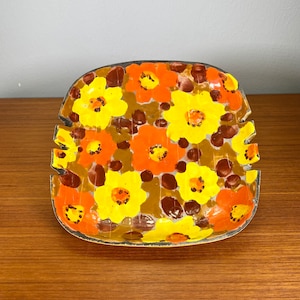 May include: A square ceramic ashtray with a floral design. The ashtray is decorated with yellow, orange, and brown flowers on a brown background.