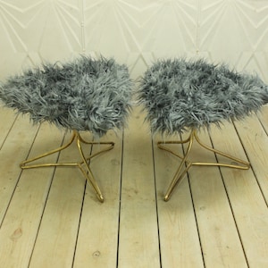 May include: Two gray faux fur ottomans with gold metal star-shaped bases. The ottomans are sitting on a light wood floor.