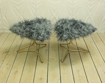 May include: Two gray faux fur ottomans with gold metal star-shaped bases. The ottomans are sitting on a light wood floor.