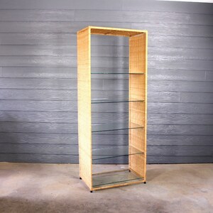 May include: A tall, woven wicker bookshelf with five glass shelves. The bookshelf has a natural wood color and is standing on a concrete floor.
