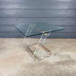 May include: A clear acrylic table with a glass top and a brass rod accent. The table has a unique X-shaped base.