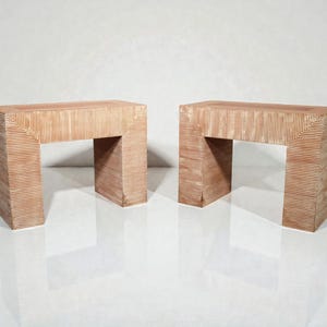 May include: Two light brown, rectangular side tables with a unique architectural design. Each table features a horizontal top supported by two vertical legs, creating an open space. The tables have a textured, ridged surface.