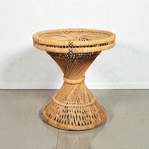 May include: A natural woven wicker side table. The round tabletop is supported by a flared, hourglass-shaped base. The table is made of light brown wicker and has a decorative woven pattern.