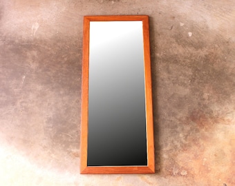 May include: A rectangular mirror with a wooden frame. The frame is a light brown color. The mirror is hanging on a textured wall.