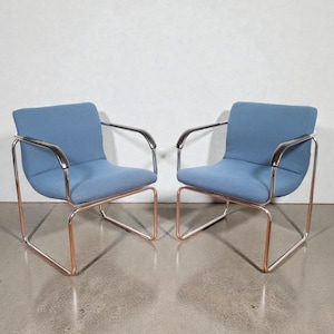 May include: Two modern armchairs with a light blue fabric seat and backrest. The chairs have a chrome frame with copper-colored legs. The armrests are also chrome. The chairs are placed against a white wall.