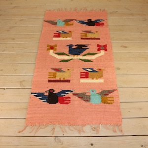 May include: A pink woven rug with a geometric pattern of stylized birds and flowers. The rug has a fringe border.