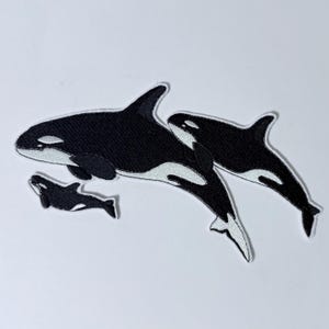 Könnte beinhalten: Drei gestickte Orca-Wal-Patches in Schwarz und Weiß. Der größte Orca schwimmt über den beiden anderen, die unter ihm schwimmen.