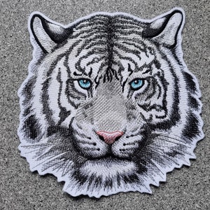Könnte beinhalten: Gestickter Aufnäher mit dem Kopf eines weißen Tigers mit blauen Augen. Der Aufnäher ist mit schwarzen und weißen Stickereien und einer rosa Nase detailliert.