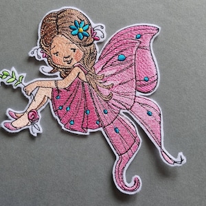 Könnte beinhalten: Gestickter Aufnäher mit einer sitzenden Fee mit braunen Haaren und rosa Flügeln. Die Fee trägt ein rosa Kleid und blaue Akzente auf ihren Flügeln. Sie hält einen Zweig mit Blättern und eine Rose.