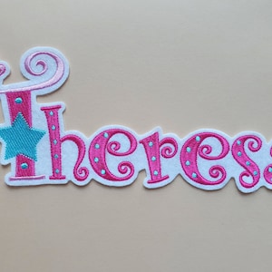 Könnte beinhalten: Rosa und blauer gestickter Aufnäher mit dem Namen "Theresa" in einer verspielten Schriftart. Der Aufnäher zeigt einen kleinen Schmetterling und einen Stern.