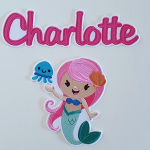 Könnte beinhalten: Gestickter Aufnäher mit einer Cartoon-Meerjungfrau mit rosa Haaren, einem blauen Bikini und einem türkisfarbenen Schwanz, die eine blaue Qualle hält. Der Name "Charlotte" ist oben in rosa Schrift gestickt.