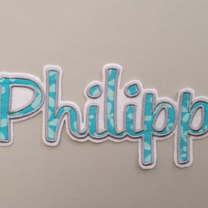 Könnte beinhalten: Ein türkisfarbener und grauer Stoff-Patch mit dem Namen "Philipp", der in weißer Schrift gestickt ist.
