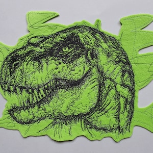Könnte beinhalten: Ein grüner Filzfleck mit einem schwarzen gestickten Bild eines Dinosaurierkopfes. Das Auge des Dinosauriers ist sichtbar und der Mund ist offen, so dass die Zähne zu sehen sind.