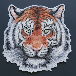 Könnte beinhalten: Nahaufnahme eines gestickten Tigergesichts-Patches mit orangefarbenen, weißen und schwarzen Streifen. Der Tiger hat grüne Augen und einen grimmigen Ausdruck.