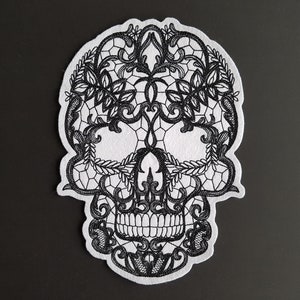 Könnte beinhalten: Schwarz-weißes Totenkopf-Design aus Spitze. Der Totenkopf besteht aus aufwendiger Spitze mit floralen Mustern.