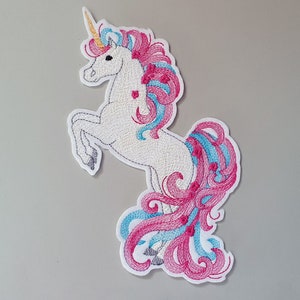 Könnte beinhalten: Gestickter Aufnäher mit einem weißen Einhorn mit rosafarbenem und blauem Mähne und Schweif. Das Einhorn steht auf seinen Hinterbeinen und hat den Kopf zur Seite gedreht.