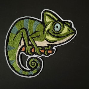 Könnte beinhalten: Grüne und blaue gestickte Chamäleon-Aufnäher mit weißem Umriss. Das Chamäleon ist zusammengerollt, mit dem Schwanz um den Körper gewickelt.