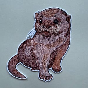 Könnte beinhalten: Gestickter brauner und weißer Otter-Patch. Der Otter sitzt mit seinen Pfoten unter dem Körper und seinem Schwanz hinter sich eingerollt.