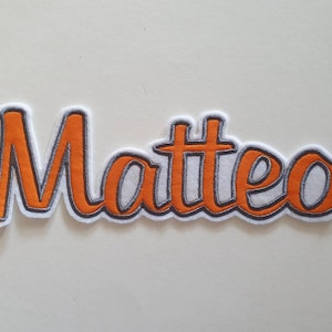 Könnte beinhalten: Ein orangefarbener, gestickter Aufnäher mit dem Namen "Matteo" in Schreibschrift. Die Buchstaben sind schwarz-weiß umrandet, mit weißem Hintergrund. Der Aufnäher ist wahrscheinlich für Kleidung oder Accessoires.