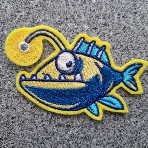 Könnte beinhalten: Ein gestickter Aufnäher in Gelb und Blau, der einen Cartoon-Anglerfisch mit einem leuchtenden Köder zeigt. Der Anglerfisch hat ein großes, rundes Auge und ein zahniges Grinsen.