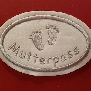 Könnte beinhalten: Weißer ovaler Stoff-Patch mit silberner Stickerei von zwei Baby-Fußabdrücken und dem Wort "Mutterpass".